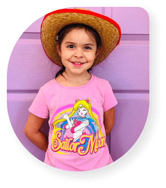 Cheerful preschool girl in Las Vegas wearing a pink Sailor Moon shirt and cowboy hat, standing against a purple wall.